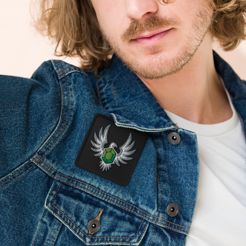 Elven Stone embroidered patch with a green gemstone framed by silver wings, worn on a blue denim jacket.
