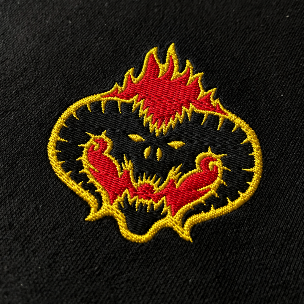 Close-up of a horned face outlined in red and gold flames embroidered on black fabric, showing the Fire Demon hoodie design.