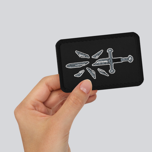 Hand holding the The Broken Blade embroidered patch, showing a silver broken sword and scattered blade fragments and the stitched border.