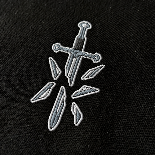 Close-up of a silver broken sword and scattered blade fragments embroidered on black fabric, showing the The Broken Blade T-shirt design.